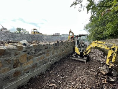 Mini excavator working alongside a long stone retaining wall under cloudy skies