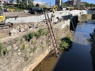 Workers repointing a tall stone retaining wall above a canal using extension ladders