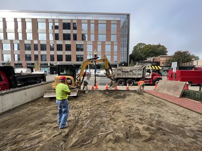 Compact excavator and dump truck preparing graded subgrade for sidewalk pour downtown