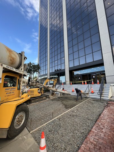 Concrete mixer truck discharging chute as worker screeds sidewalk pour beside high-rise