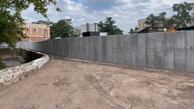 Long poured concrete retaining wall stands beside dirt yard with stone wall opposite stream