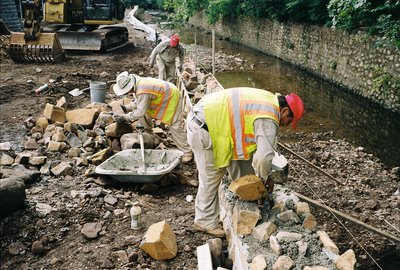 Masons rebuild stone retaining wall along streambed with excavator and wheelbarrow on site