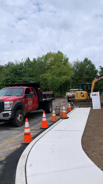 Red dump truck and excavator beside fresh concrete sidewalk pour with traffic cones