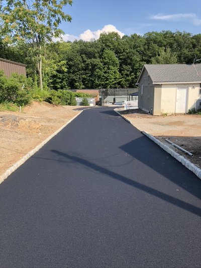 Freshly paved asphalt driveway with cobblestone curbing leading toward a small outbuilding under blue sky