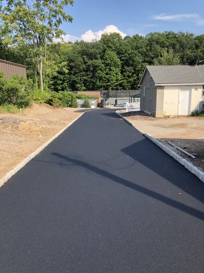 Newly paved asphalt drive with stone curbing curving past a residential outbuilding on sunny day