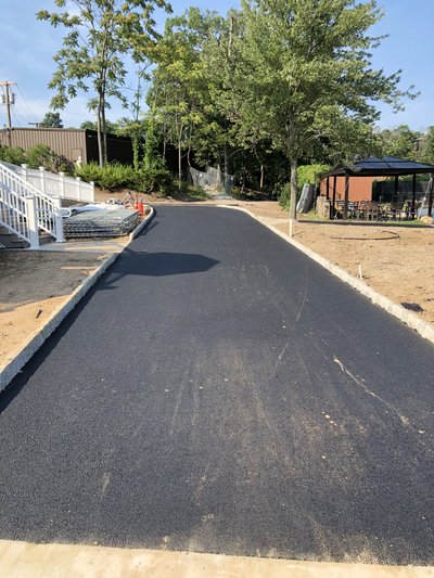 Freshly laid asphalt path bordered by stone curbing leading toward a tree-shaded patio area
