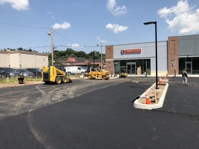 Skid-steer and roller working on a freshly paved commercial parking lot beside a retail building