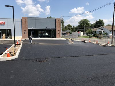 Workers raking and finishing fresh asphalt in a commercial parking lot beside a retail storefront