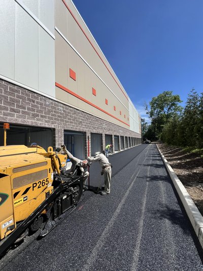 Yellow asphalt paver laying fresh mat alongside a large commercial warehouse on a sunny day