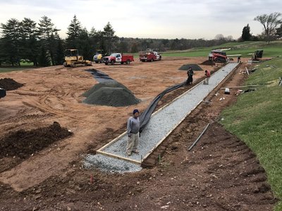 Worker beside gravel base in formed cart path trench with bulldozers and gravel piles