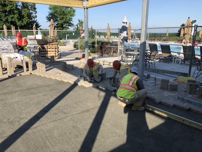 Workers in safety vests laying paver patio beside fenced swimming pool deck