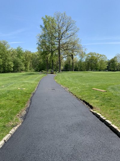 Newly paved asphalt cart path curving through manicured fairway and wooded grounds in sunshine