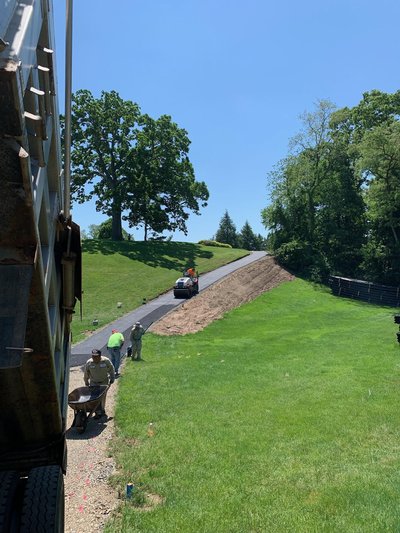 Crew paving asphalt cart path on grassy hillside beside dump truck under blue sky